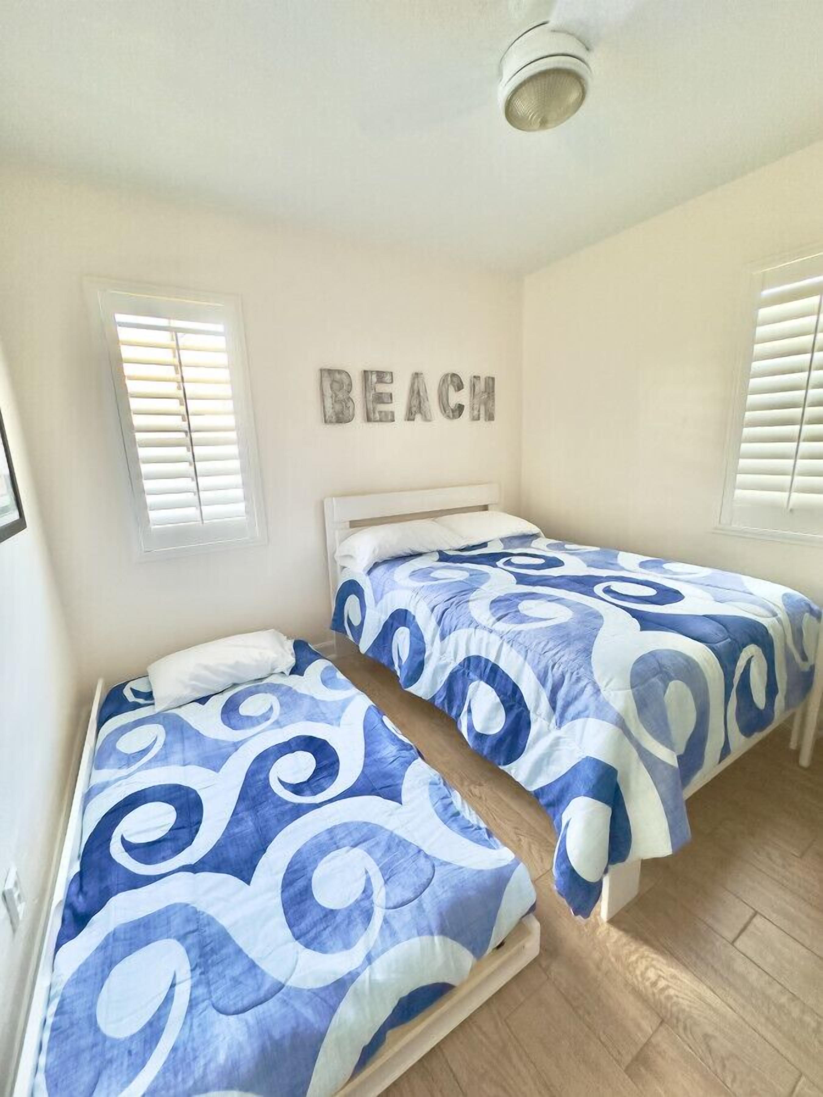 Een slaapkamer met twee bedden, een plafondventilator en de tekst 'BEACH' op de muur.