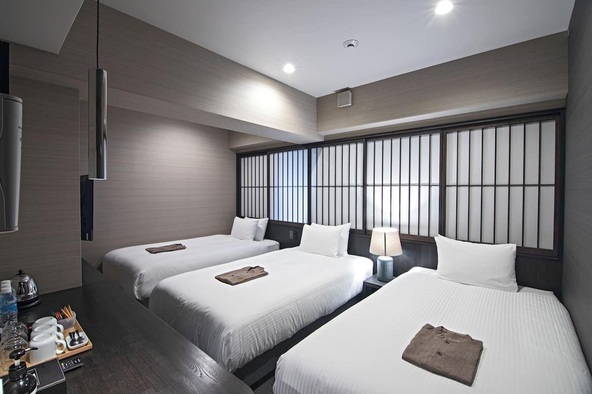 A hotel room with three beds, a minibar, a TV, and a large window with blinds.
