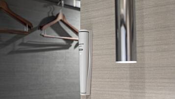 A wall-mounted device, possibly a thermostat or control panel, in a room with a textured wall and a wooden hanger in the background.