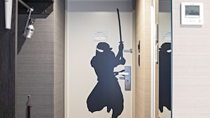 A room with a door featuring a silhouette of a samurai holding a sword, an emergency exit sign, and a wall-mounted thermostat. - HOTEL NINJA BLACK (Kyoto)
