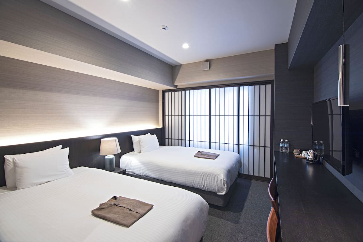 A modern hotel room with two beds, a bedside table with a lamp, a large window with blinds, and a flat-screen TV mounted on the wall.