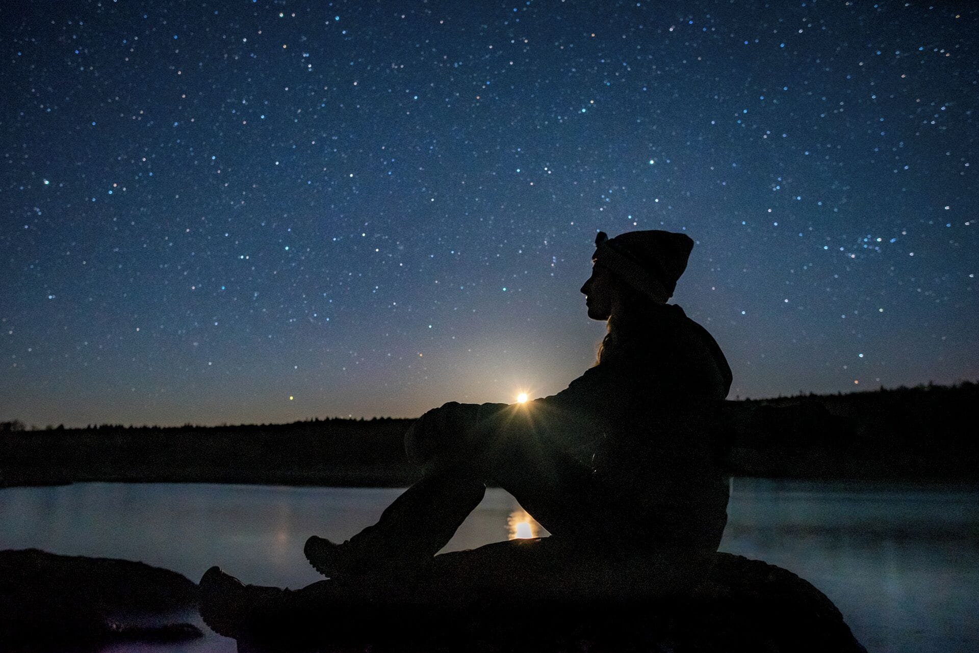 A person sitting on a rock by a lake at night, gazing at the stars.