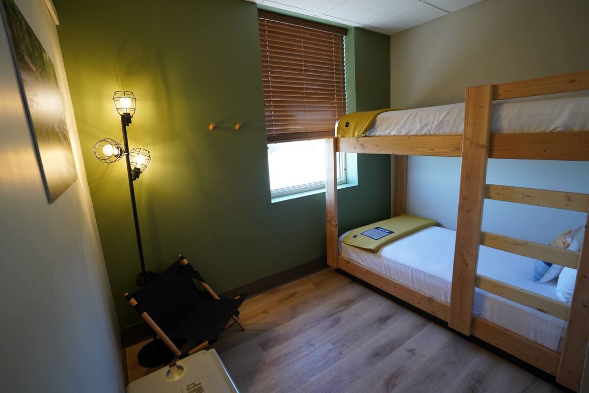 A bunk bed room with a desk and chair, a standing lamp, and a window with blinds.