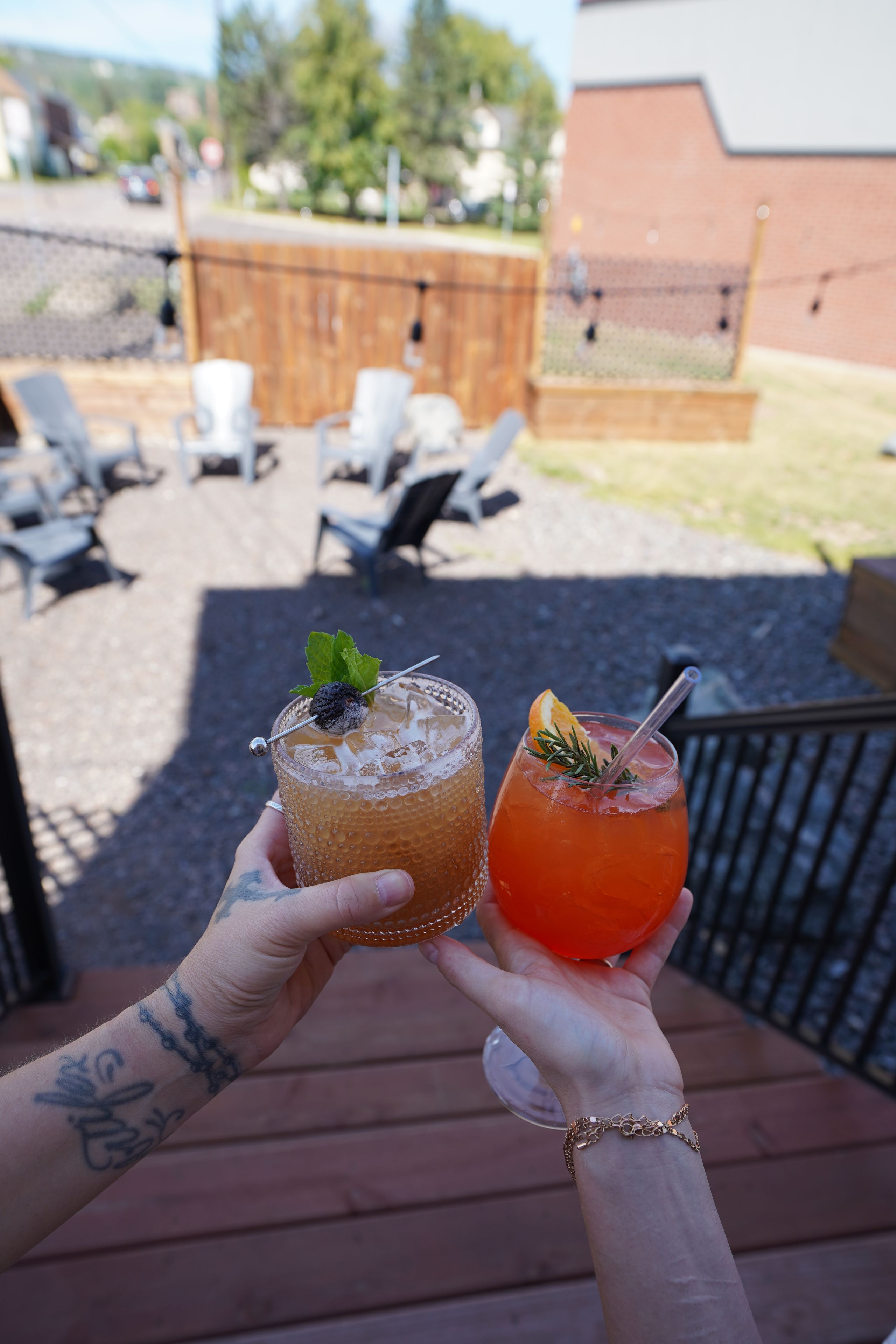 Two hands holding drinks with garnishes, a wooden deck, and outdoor seating in the background.
