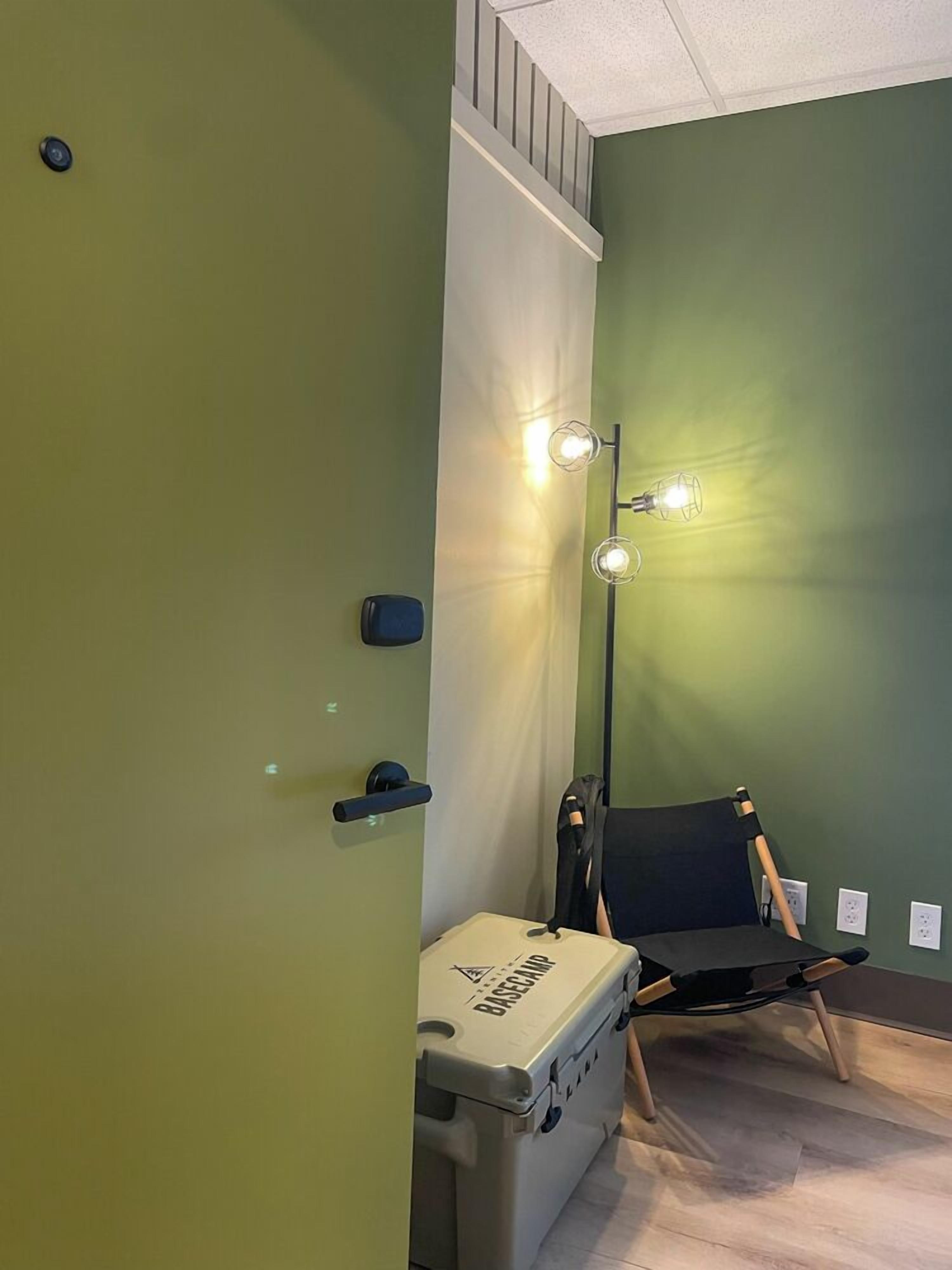 A room with green walls, a chair, a desk with a suitcase, and a floor lamp.