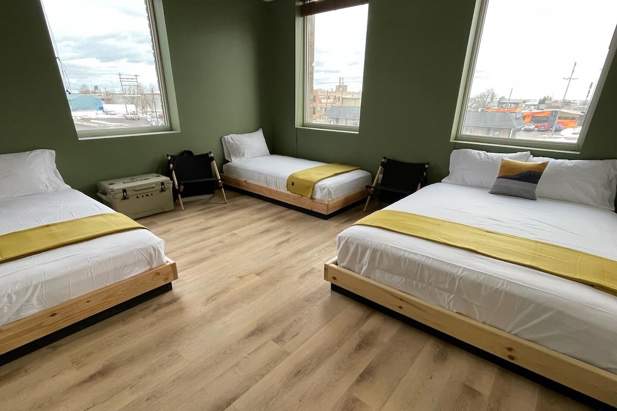 A room with three beds, each with a wooden frame and white bedding, arranged in a U-shape. There are large windows providing a view of the outside.