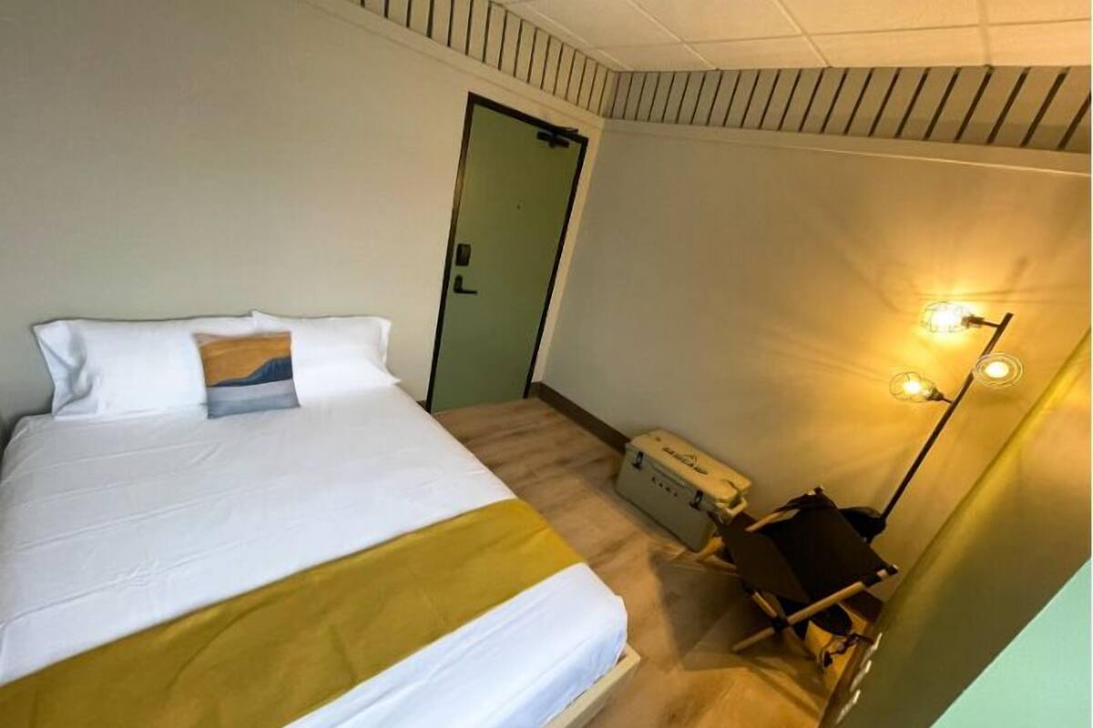 A hotel room with a bed, a desk, a chair, a suitcase, and a wall-mounted lamp.