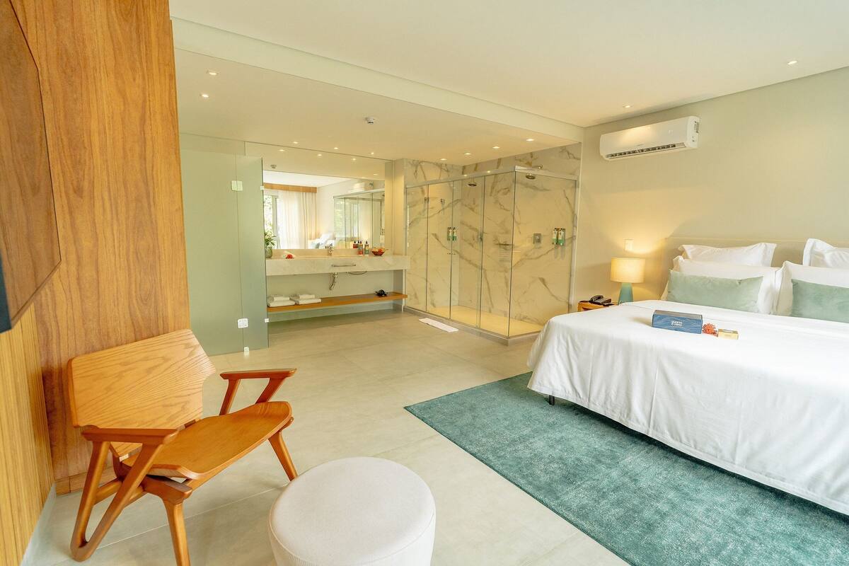 A modern hotel room with a bed, a wooden chair, a round ottoman, a bathroom with a glass shower, and a marble wall.
