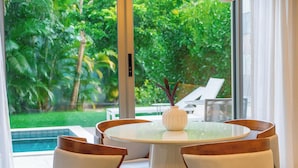 Un comedor con mesa y sillas de vidrio, un jarrón con detalles decorativos y vistas a una piscina y abundante vegetación.