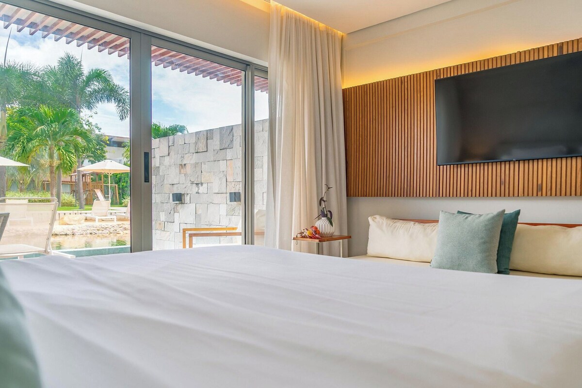 A hotel room with a large bed, a flat-screen TV mounted on a wooden panel, and a view of a pool and palm trees outside.