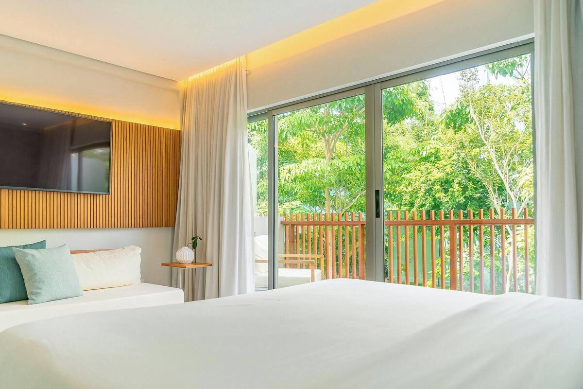 A hotel room with a bed, a TV mounted on the wall, a balcony with a view of greenery, and a wooden railing.