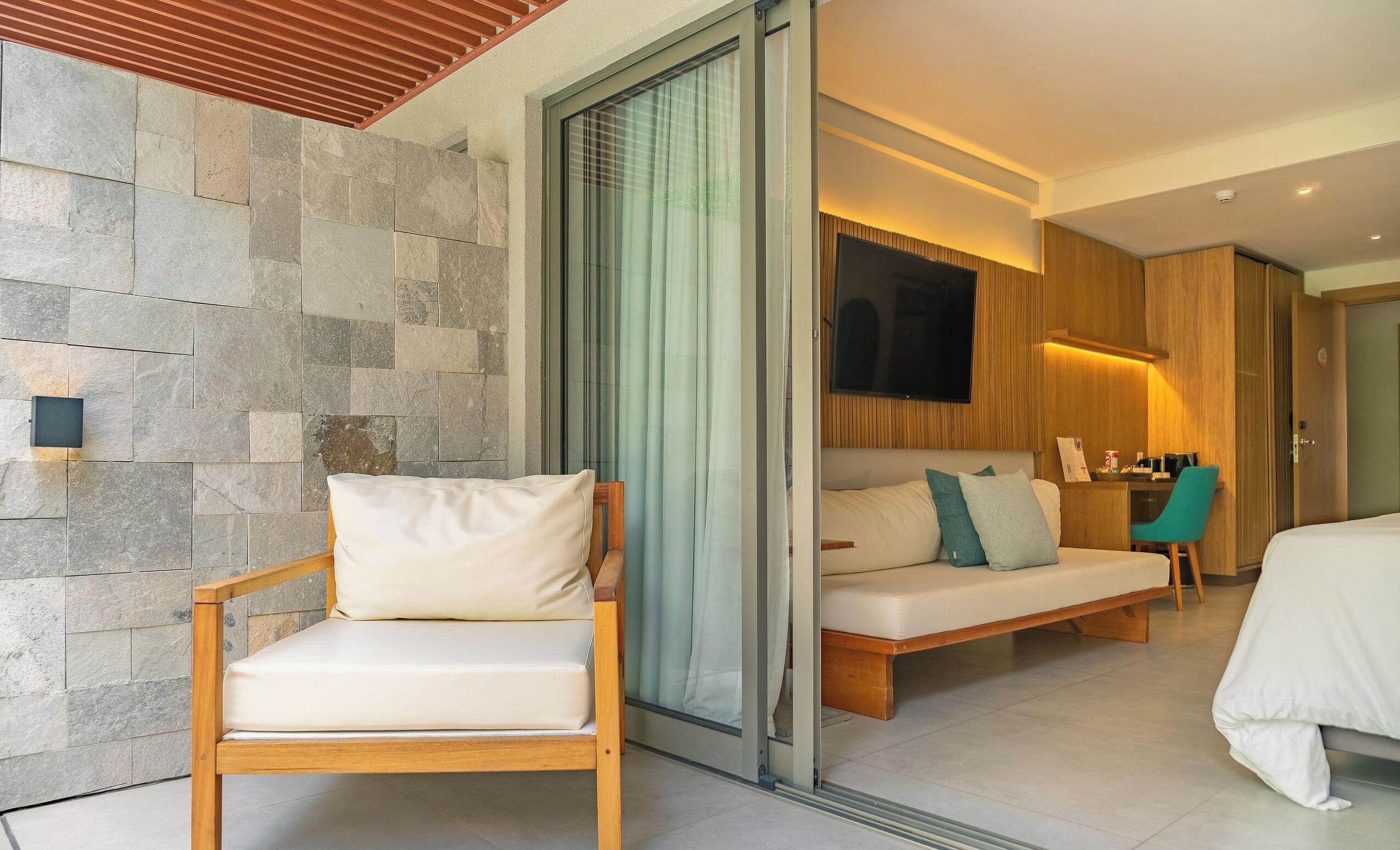 Una habitación de hotel moderna con pared de piedra, una silla de madera, un sofá con cojines, una cama y un televisor montado en la pared.