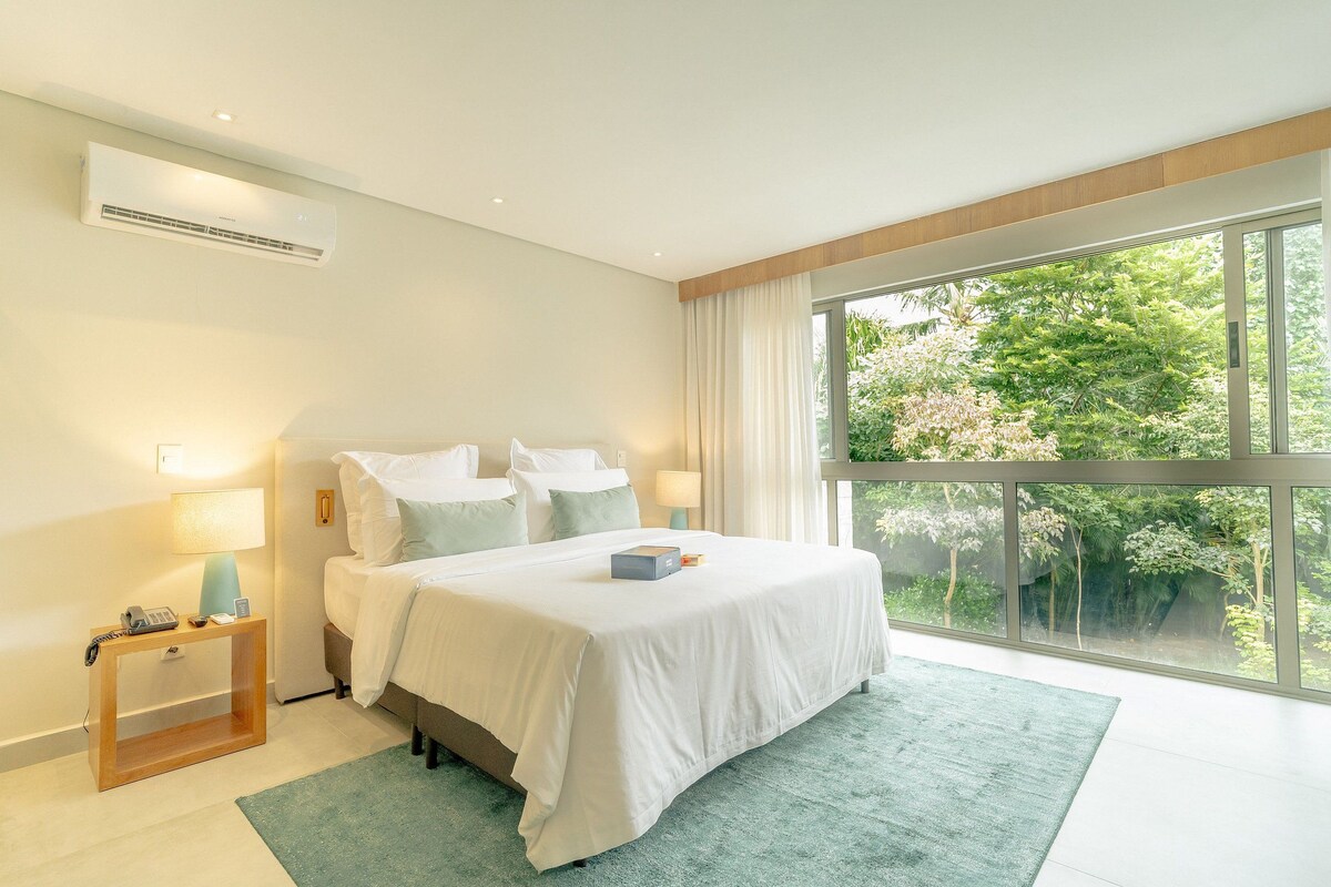 Ein modernes Hotelzimmer mit einem großen Bett, Nachttischlampen, einem Beistelltisch und Blick auf Grün durch große Fenster.