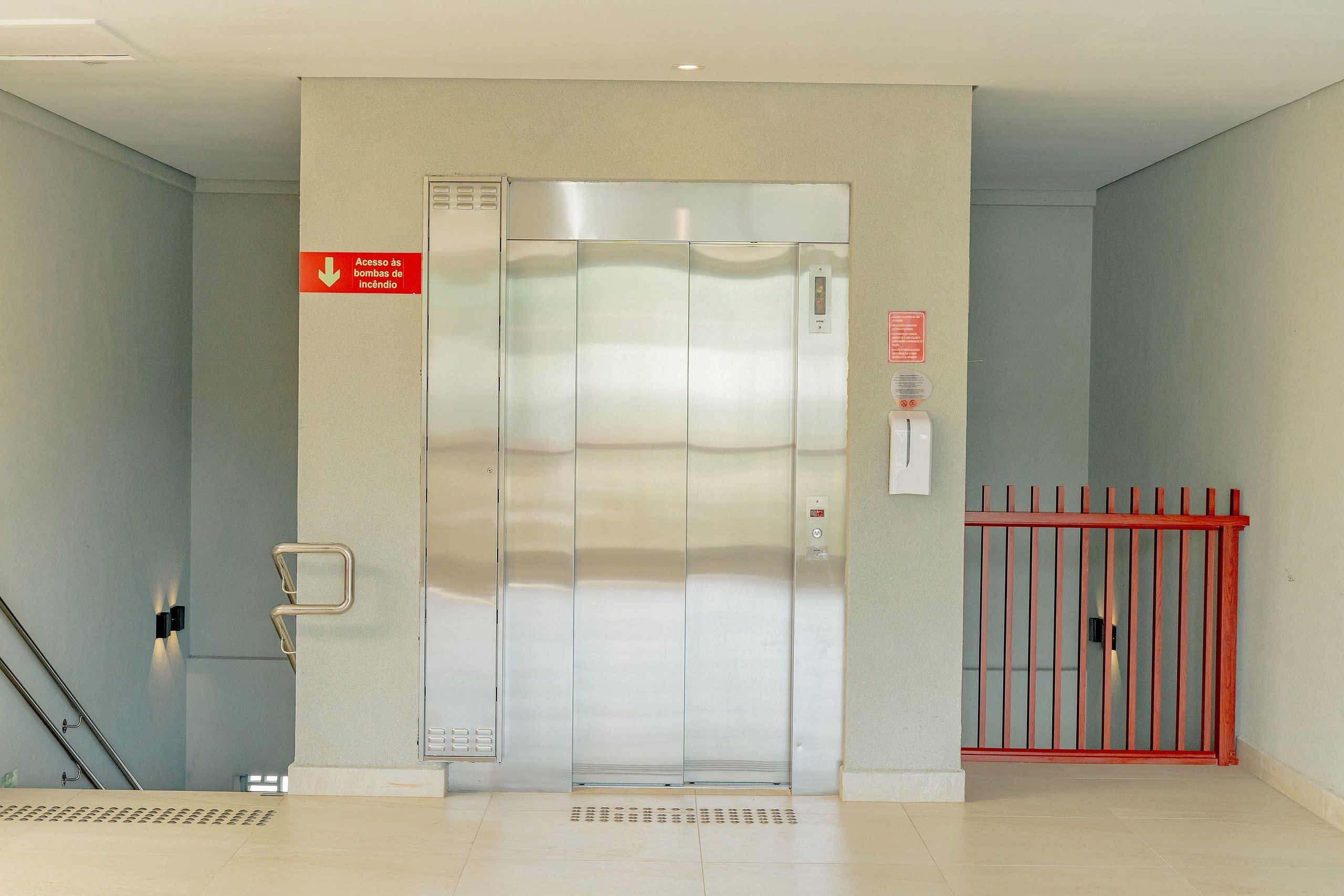 Un ascensor con acabado metálico, un letrero rojo de salida de emergencia y una puerta de seguridad roja.