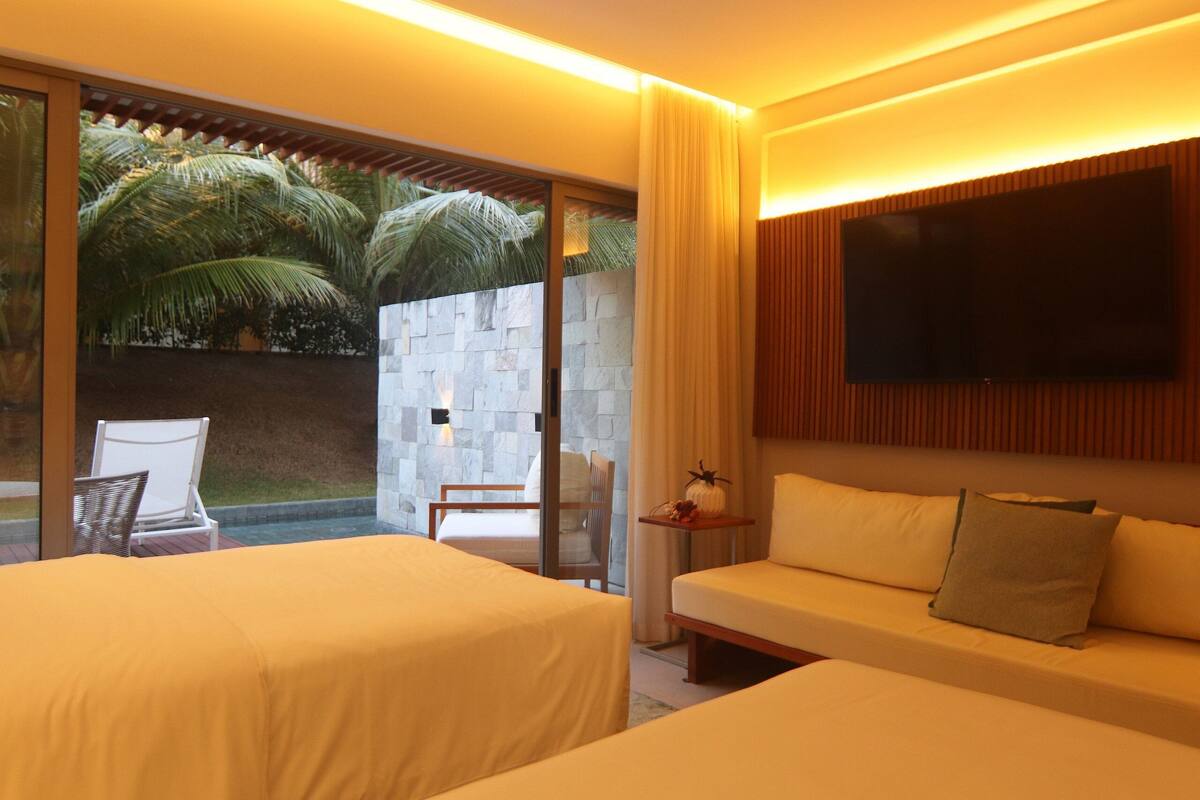 A hotel room with two beds, a TV, a sofa, a small table, and a view of palm trees.