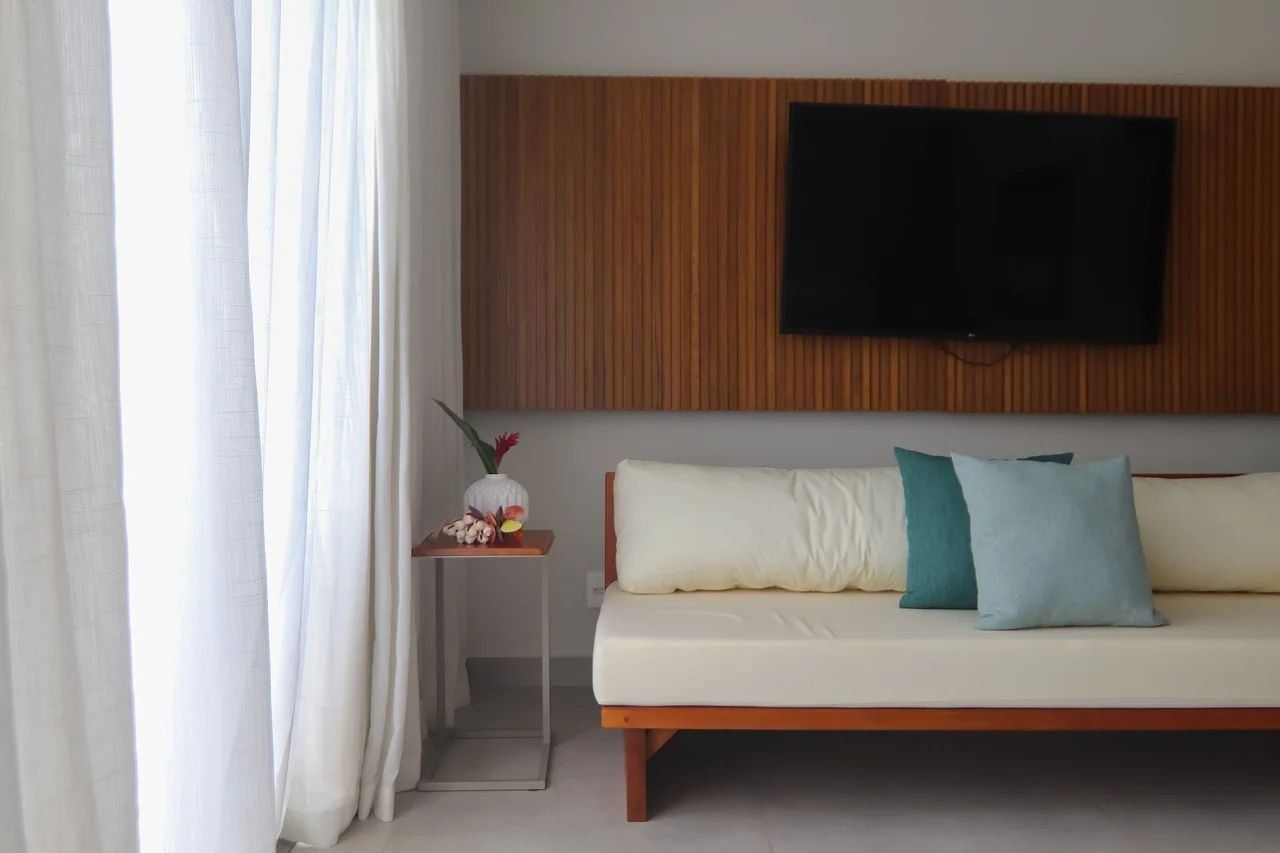 Una habitación moderna con sofá, televisor montado en una pared de paneles de madera, una mesita con un florero y cortinas de encaje.