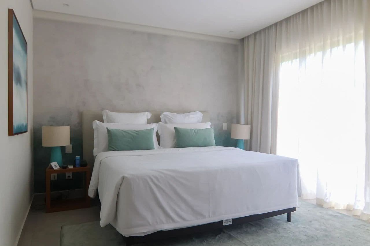 Habitación de hotel con una cama grande, dos mesitas de noche con lámparas, un cuadro enmarcado en la pared y una ventana con cortinas translúcidas.