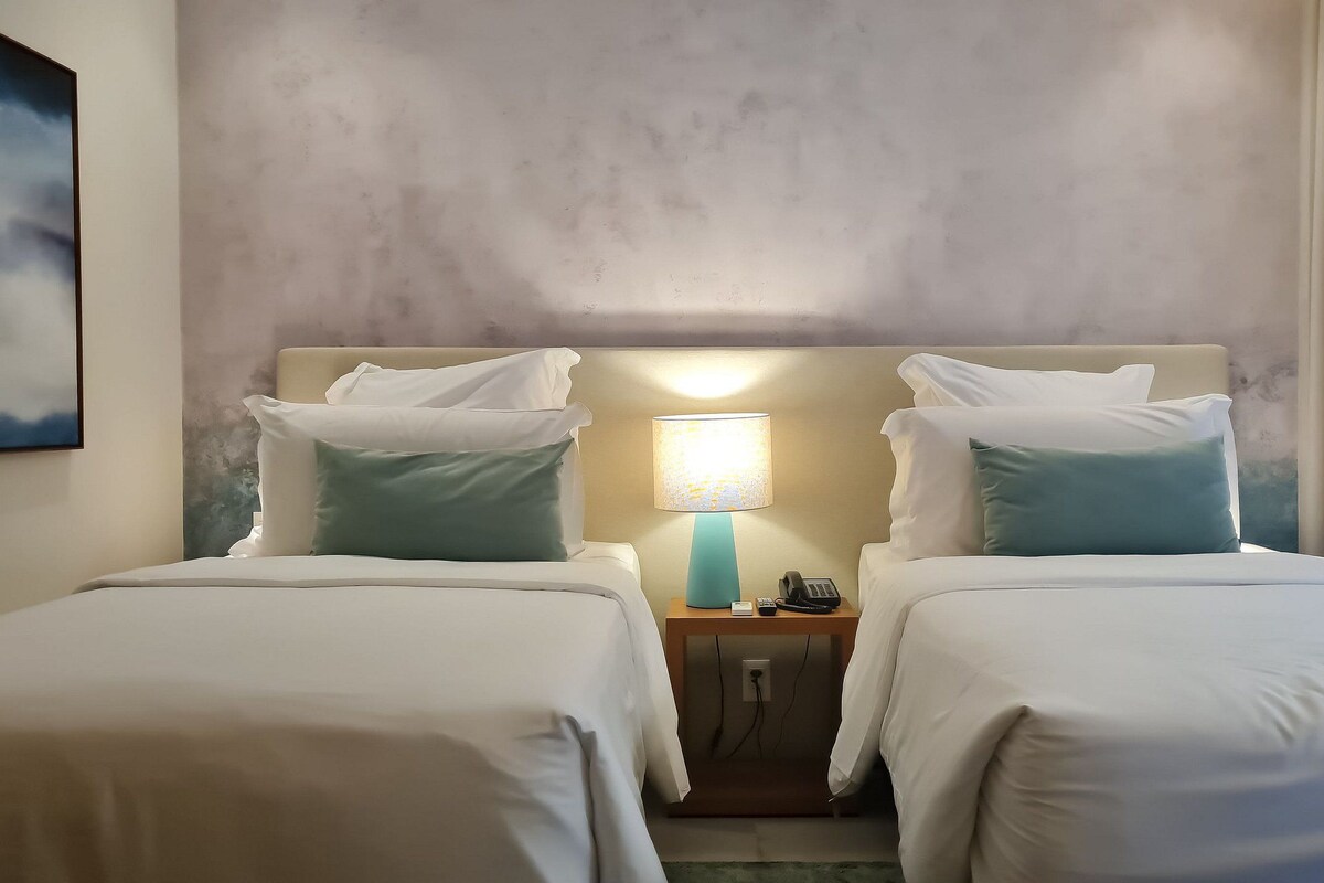 Una habitación de hotel con dos camas, una mesita de noche con lámpara, teléfono y un cuadro en la pared.