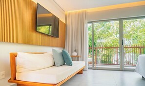 A modern hotel room with a wooden panel wall, a flat-screen TV, a sofa with cushions, a bedside table with a lamp, and a sliding glass door leading to a balcony with greenery outside. - Île de Pipa - Ma Plage Hotel (Tibau do Sul)