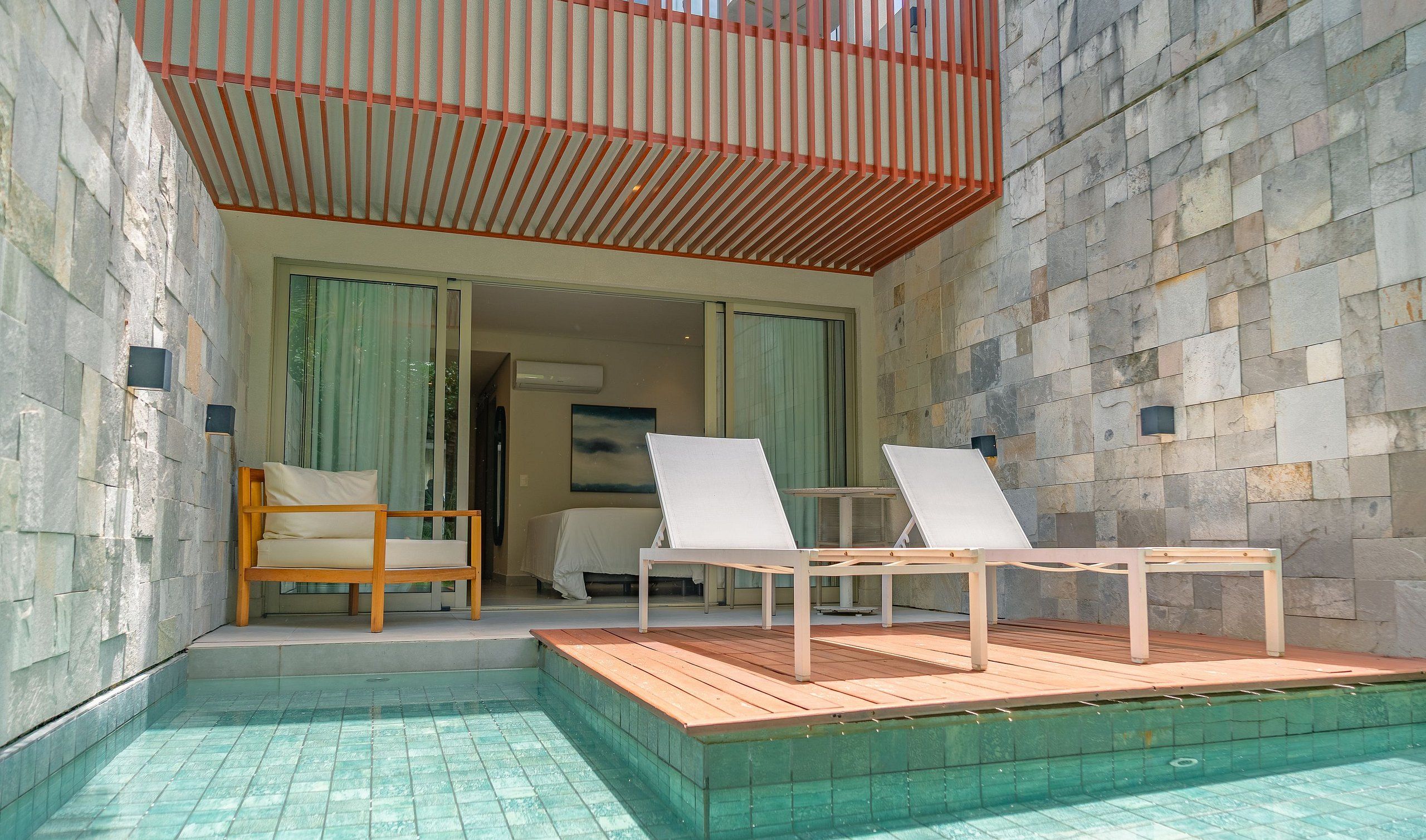 A poolside area with two lounge chairs, a wooden bench, and a view of the interior through a glass door.