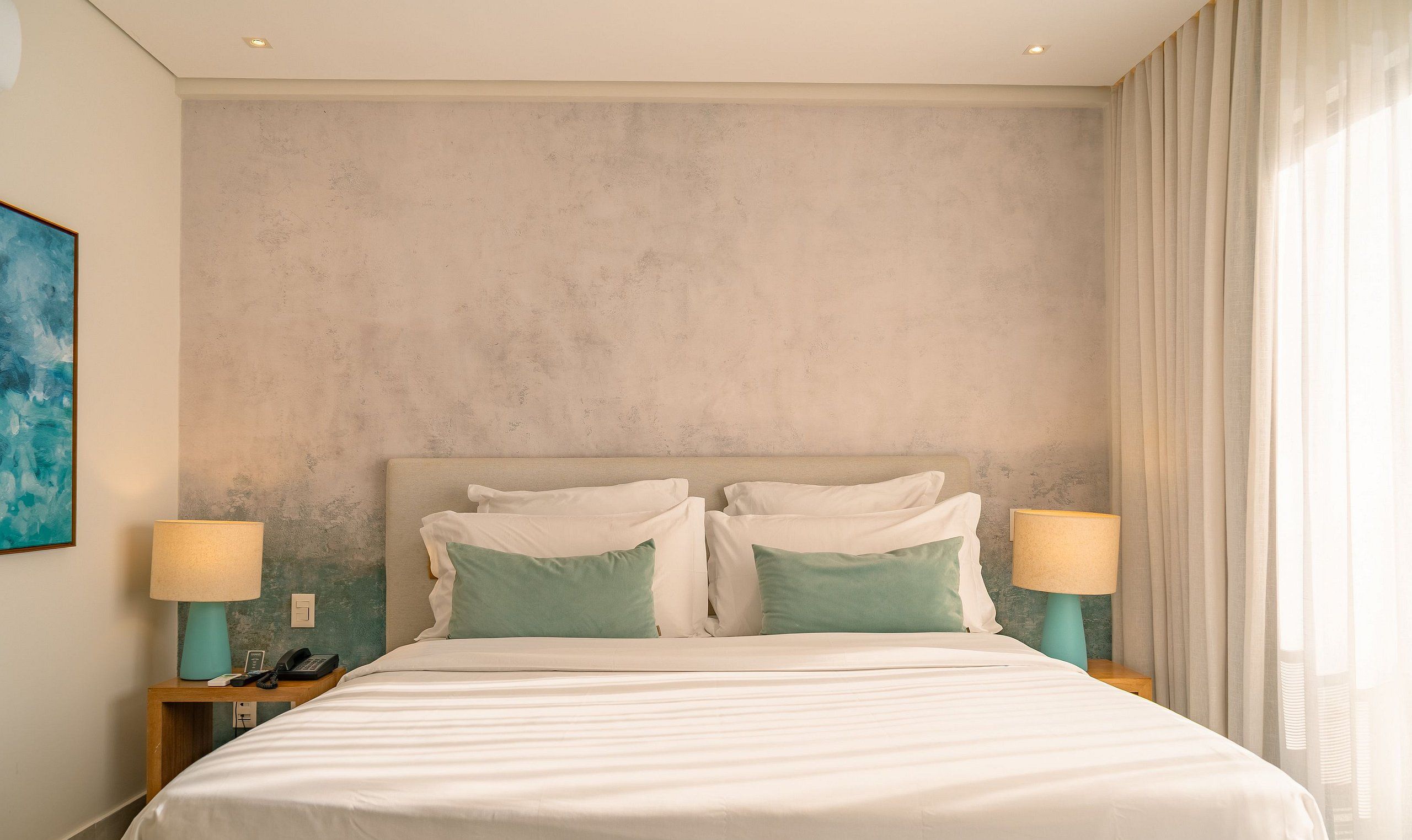 Habitación de hotel con una cama grande, dos mesitas de noche con lámparas, un cuadro en la pared y una ventana con cortinas.