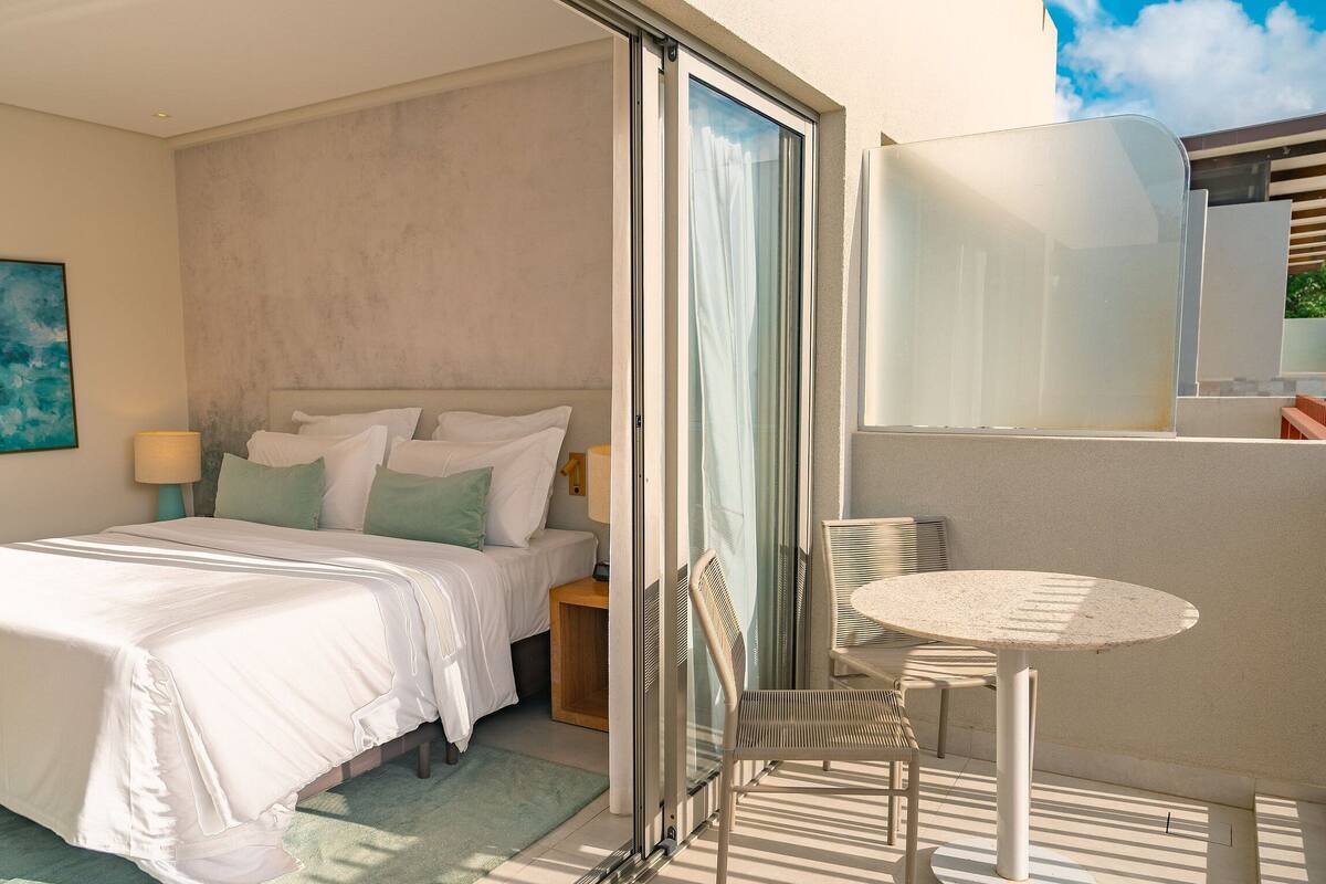 A hotel room with a bed, a small table with chairs, a window with curtains, and a painting on the wall.