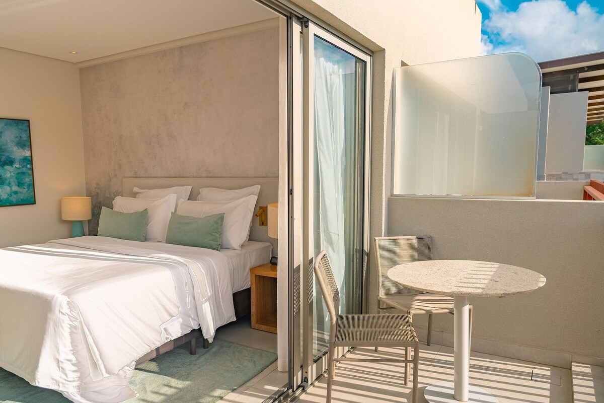 Habitación de hotel con cama, una mesita con sillas, ventana con cortinas y un cuadro en la pared.