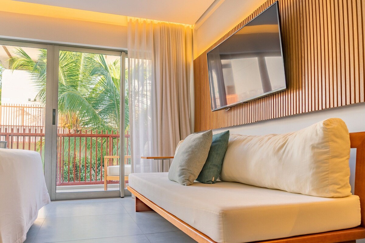 A modern hotel room with a sofa, a TV mounted on the wall, and a sliding glass door leading to a balcony with a view of palm trees.