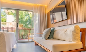 Et moderne hotelværelse med en sofa, et fjernsyn ophængt på væggen og en skydedør, der fører til en balkon med udsigt til palmer.