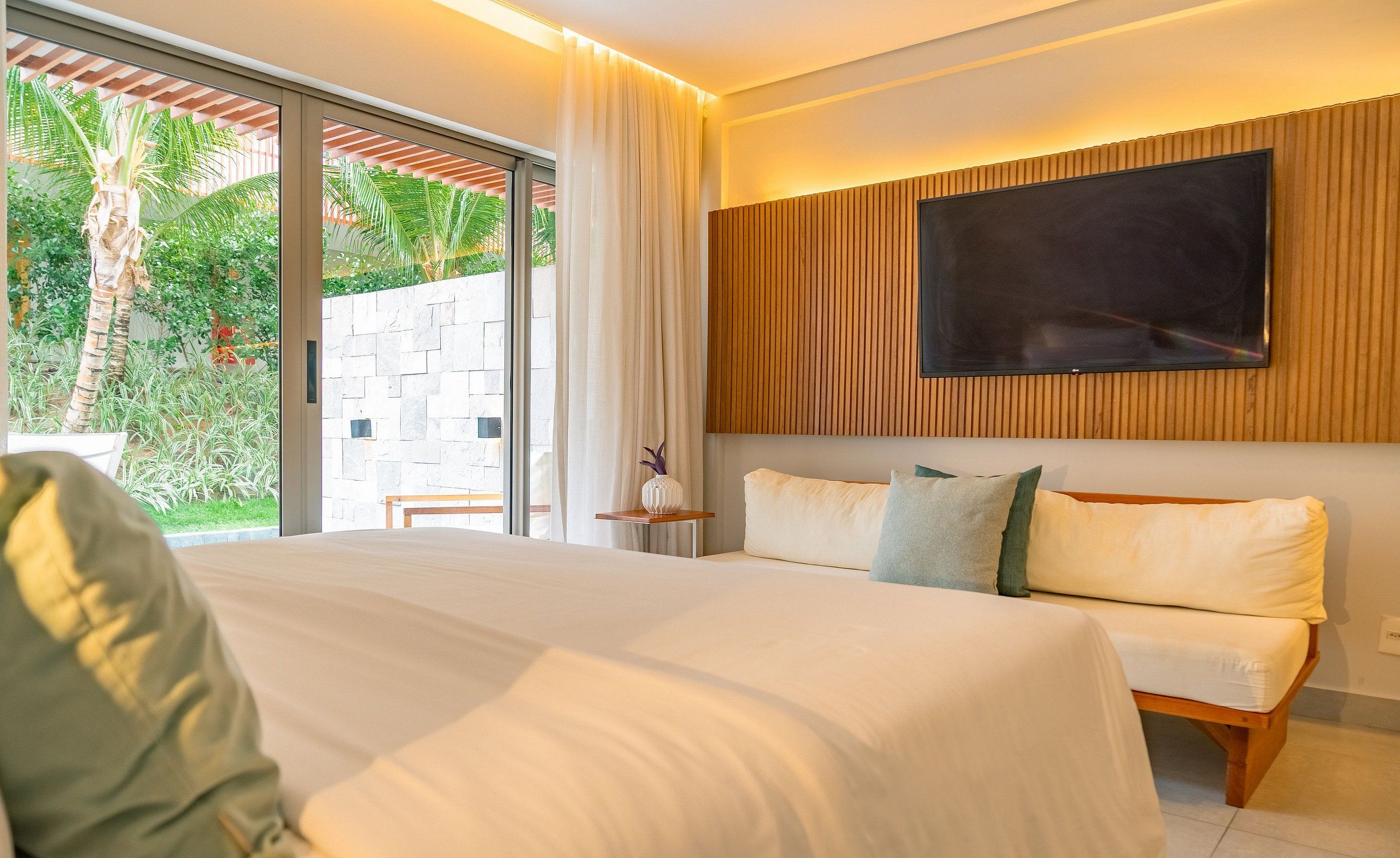 Quarto de hotel moderno com uma cama grande, uma televisão fixada na parede e vista para o verde lá fora.