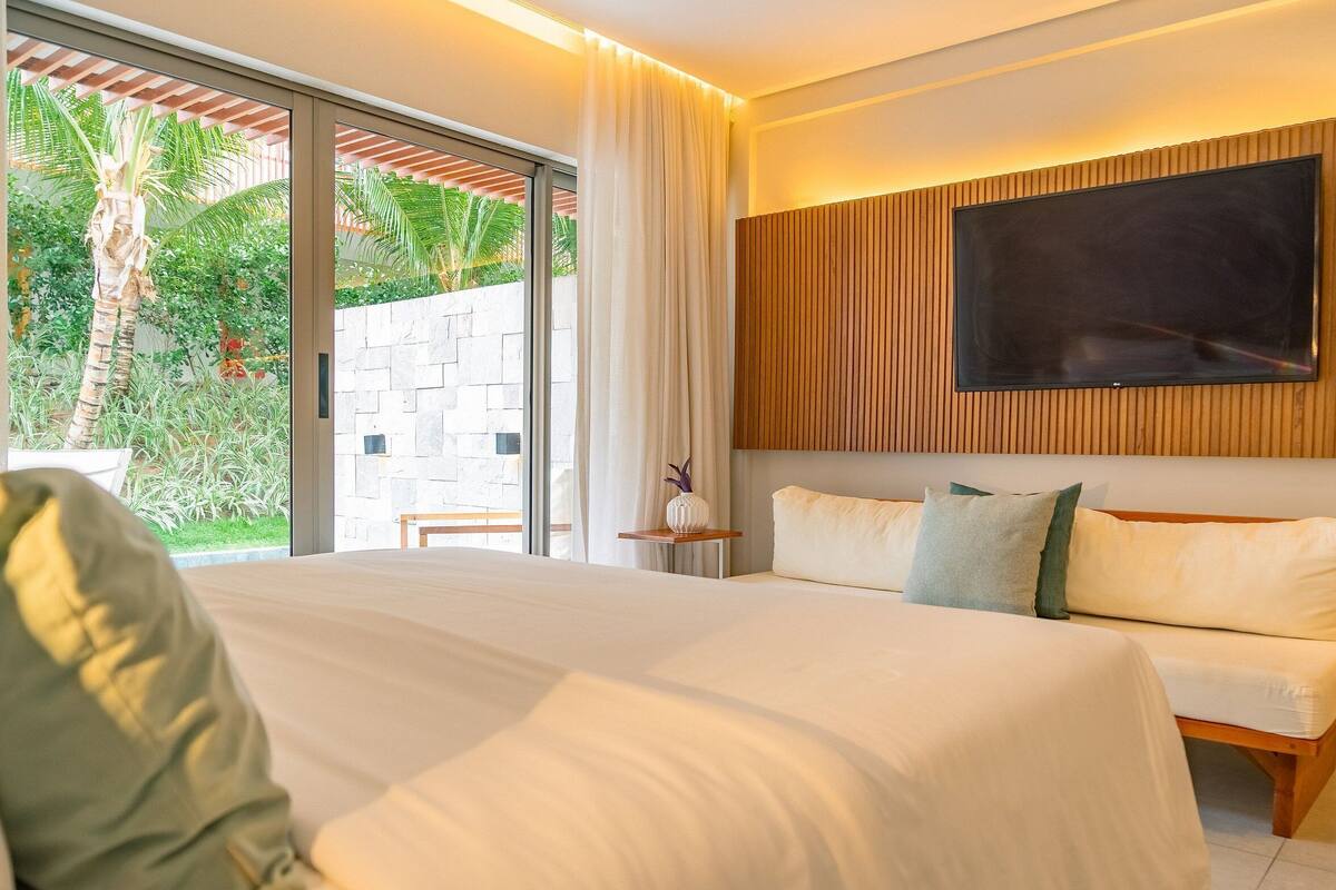 A modern hotel room with a large bed, a TV mounted on the wall, and a view of greenery outside.