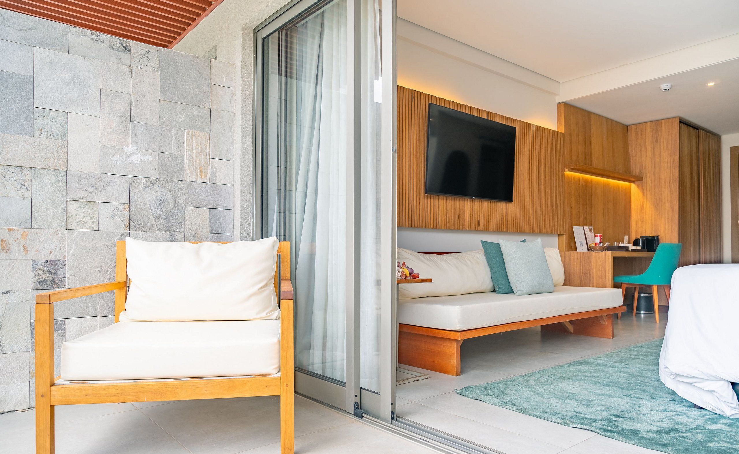 A modern hotel room with a stone wall, a wooden chair, a sofa with cushions, a TV mounted on the wall, and a small desk with a chair.