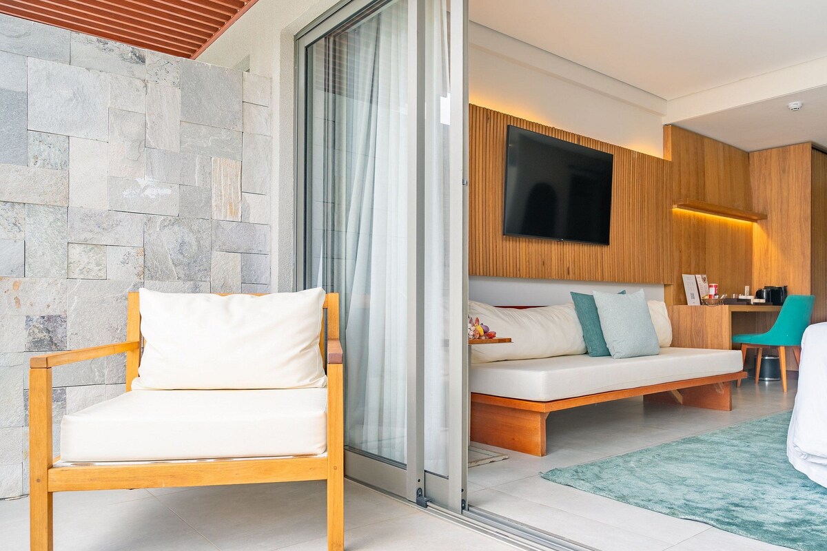 Una habitación de hotel moderna con pared de piedra, una silla de madera, un sofá con cojines, un televisor montado en la pared y un pequeño escritorio con una silla.