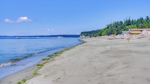 En strand med kristallklart blått vatten, en sandstrand och en skogbeklädd kulle i bakgrunden.