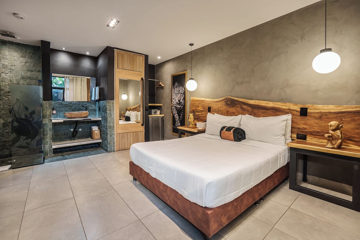 A modern bedroom with a large bed, a wooden headboard, a nightstand, a mirror, and a stone wall.