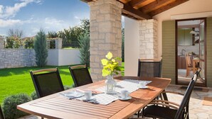 Patio esterno con tavolo e sedie in legno, un pilastro in pietra e vista su un giardino e un'area cucina.
