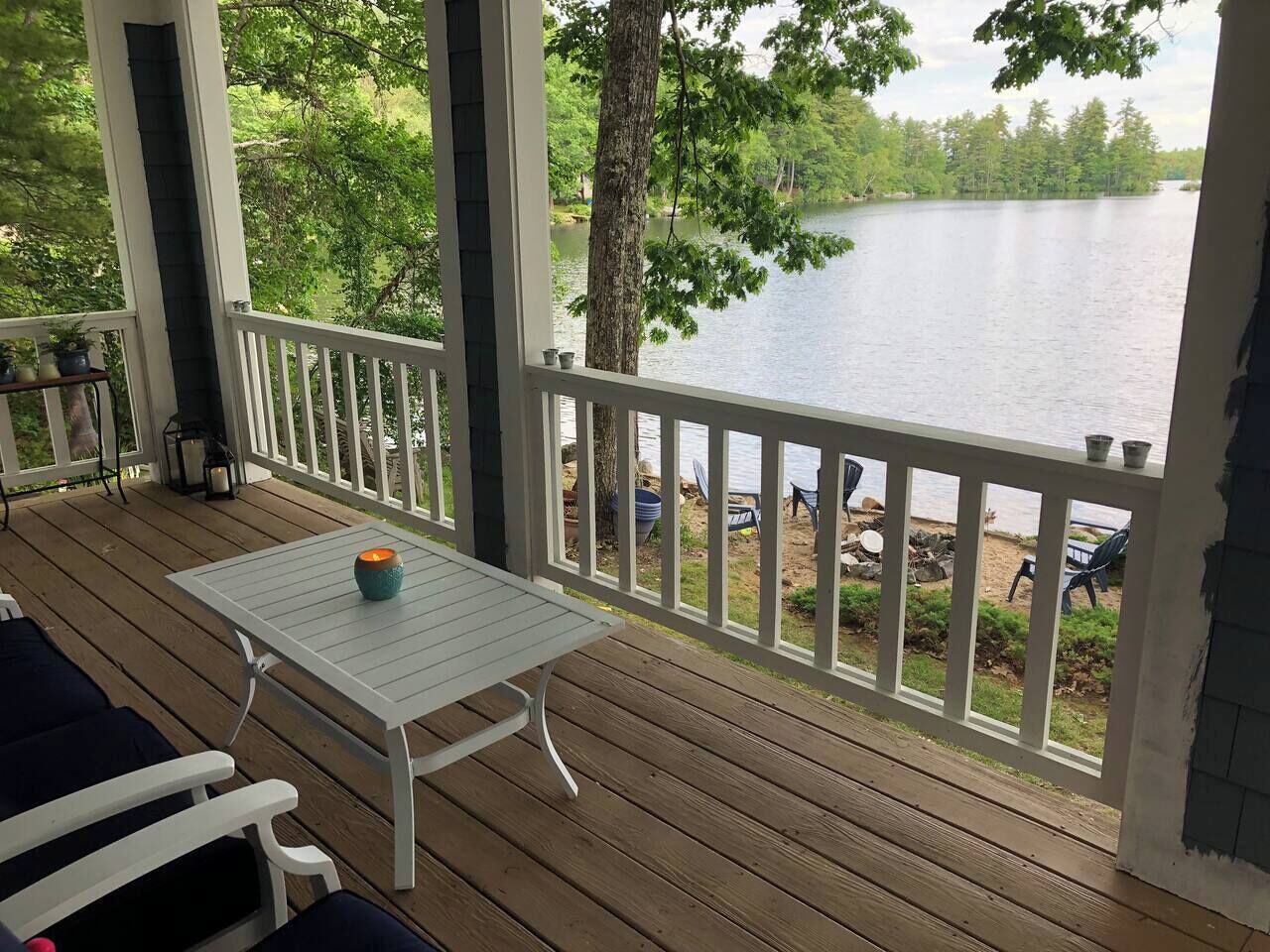 A wooden deck with a table and chairs, overlooking a lake with a dock and chairs on the shore.