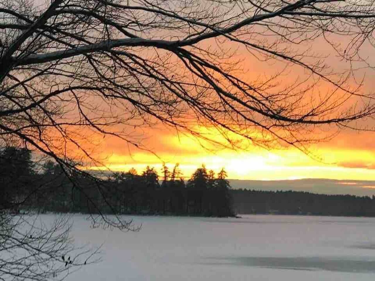 Talvinen maisema, jossa auringonlasku värjää lumipeitteisen pellon ja taustalla näkyy puita.