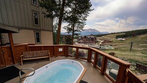 A hot tub on a wooden deck with a view of a mountain and a lodge in the background.