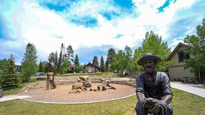 A statue of a man in a hat sitting on a stone bench in a park with a playground and a building in the background.
