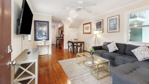 A living room with a grey sofa, a coffee table, and a TV on a stand. There is a dining area with a table and chairs, and a kitchen in the background.