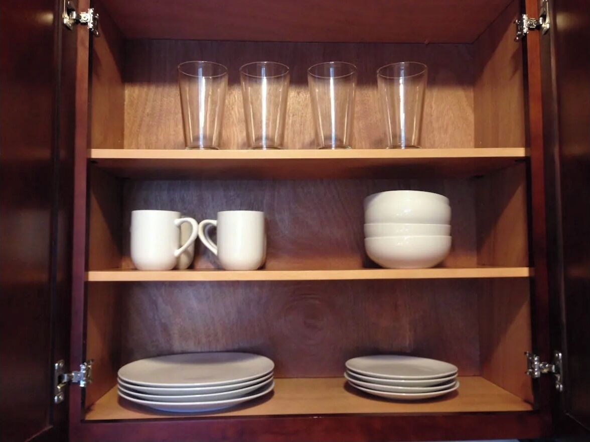 Un mueble de madera con estantes que contiene vasos transparentes, tazas blancas, cuencos blancos y platos apilados.