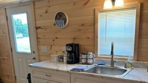 A compact kitchen with wooden walls, a sink, a coffee maker, and a window with blinds. - Between the Waters Rustic Retreat (Hilham)