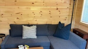 A cozy room with a wooden ceiling, a grey sofa with cushions, a small wooden table with a bowl of snacks, and a floor lamp. - Between the Waters Rustic Retreat (Hilham)