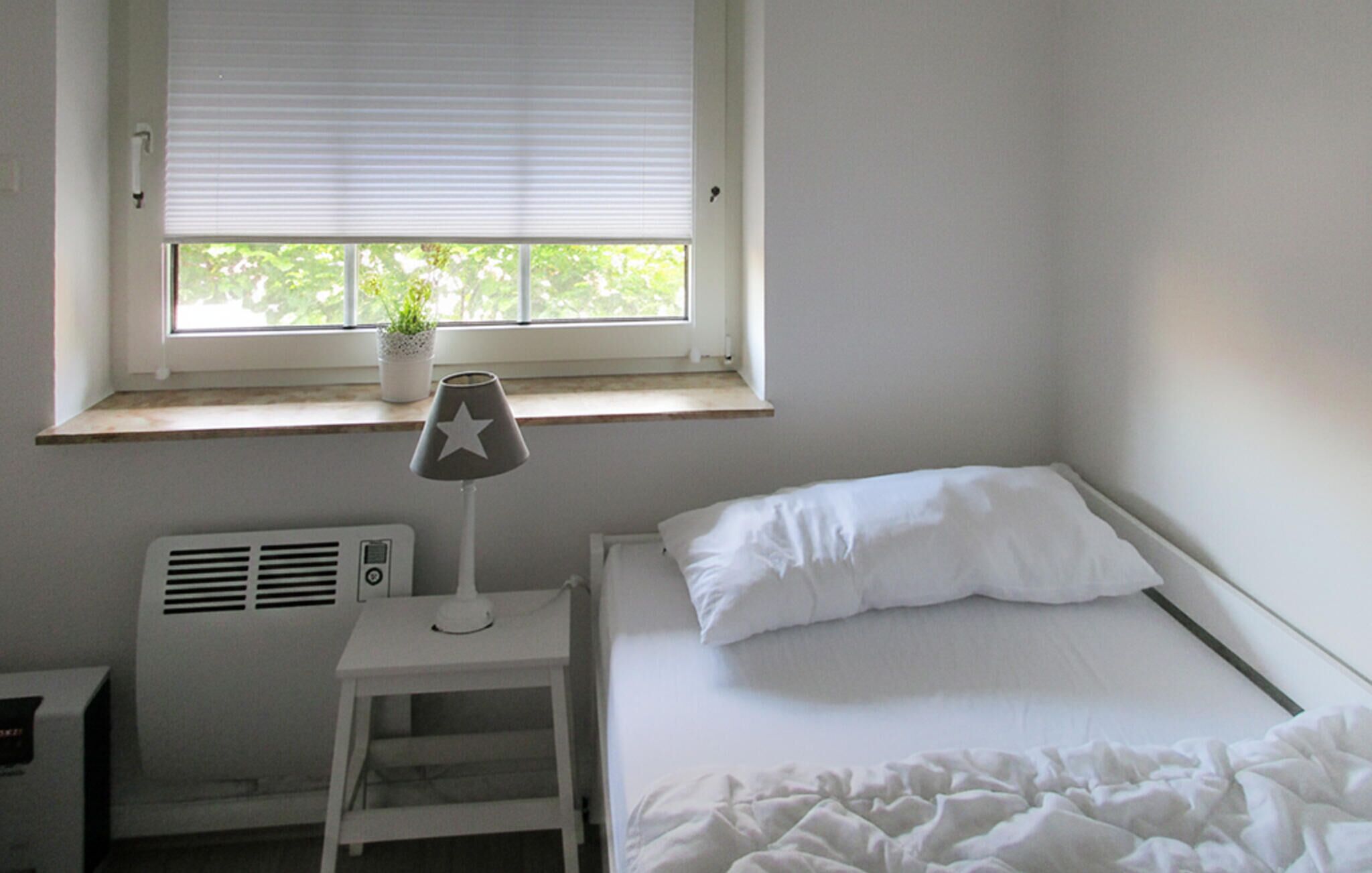A bedroom with a bed, a bedside table with a lamp, a window with blinds, and a radiator.