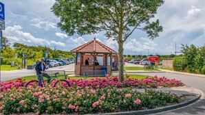 Un gazebo con tetto in tegole circondato da fiori rosa, un'area di parcheggio con auto e una persona in bicicletta.