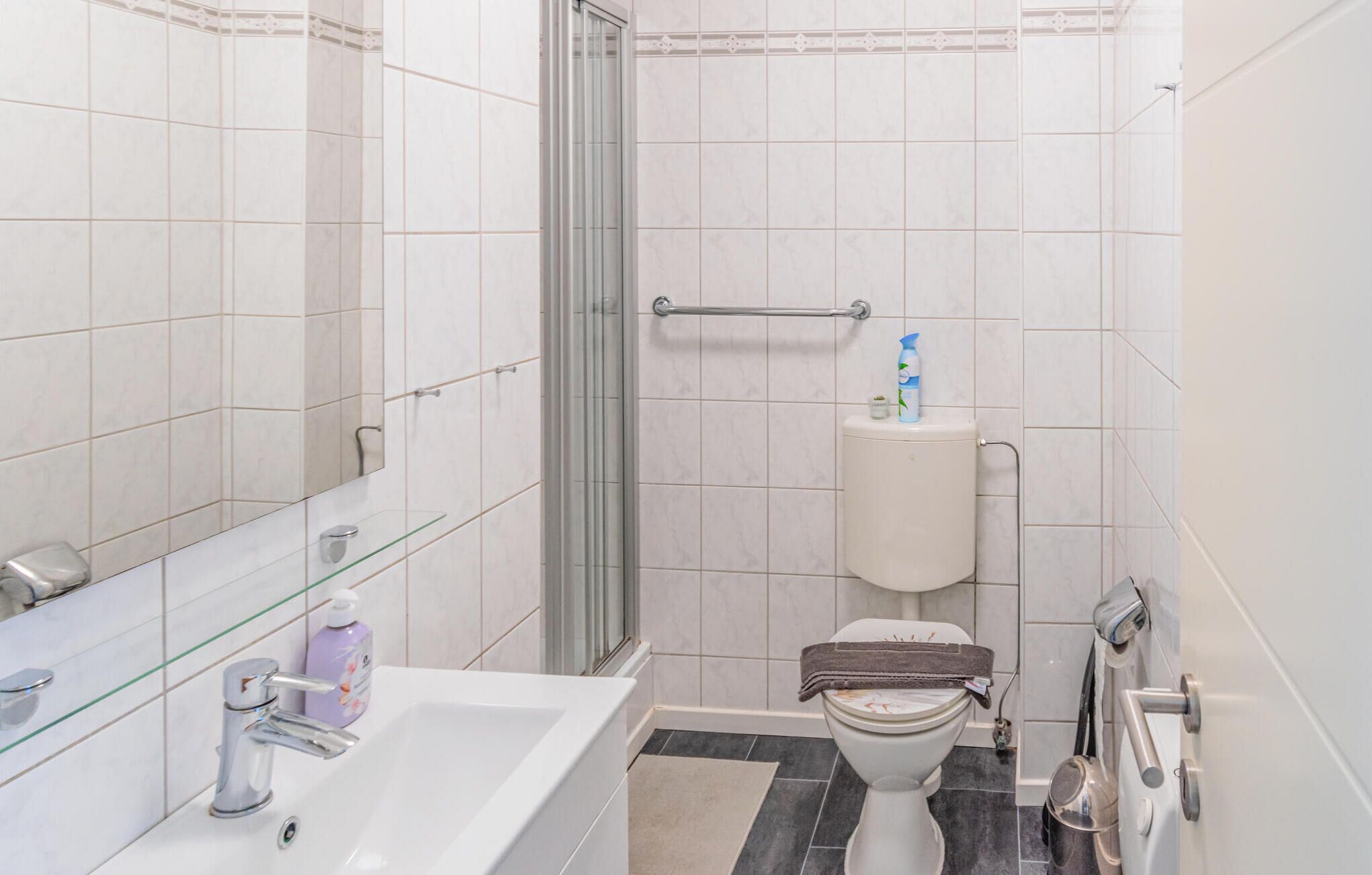 A bathroom with a glass shower enclosure, a white sink with a chrome faucet, a toilet with a towel on top, and a glass shelf with toiletries.