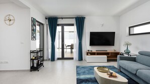 Una sala de estar moderna con una televisión de pantalla plana, un sofá, una mesa de centro con libros, un mueble para la televisión y un ventanal corredizo que da a un espacio al aire libre.