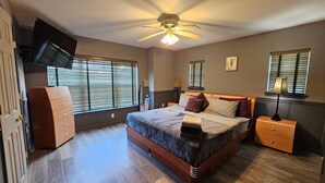Un dormitorio con cama, ventilador de techo, cómoda, televisor montado en la pared y ventanas con persianas.