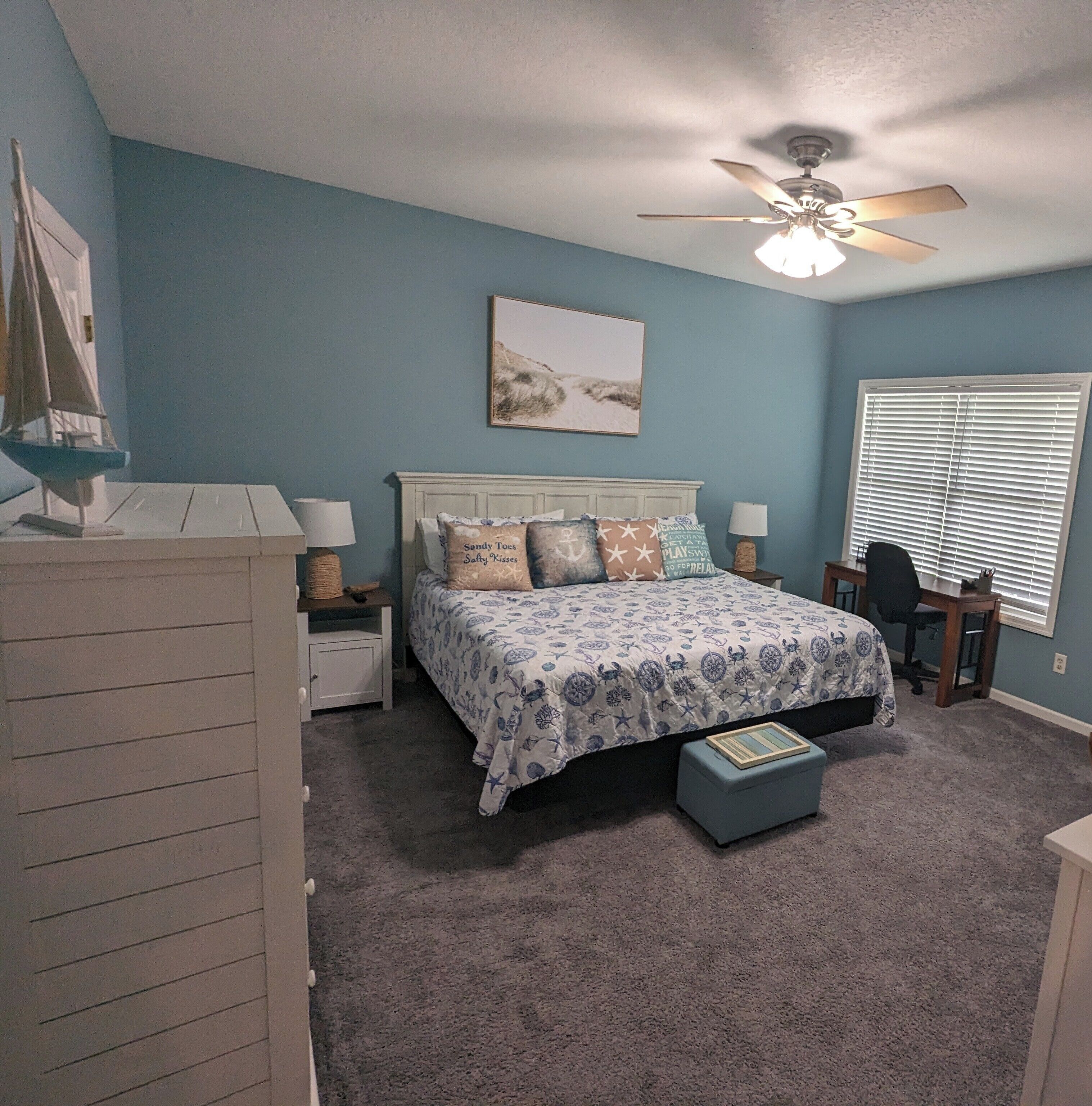 A bedroom with a bed, nightstands, a desk, a chair, a ceiling fan, and a window with blinds.